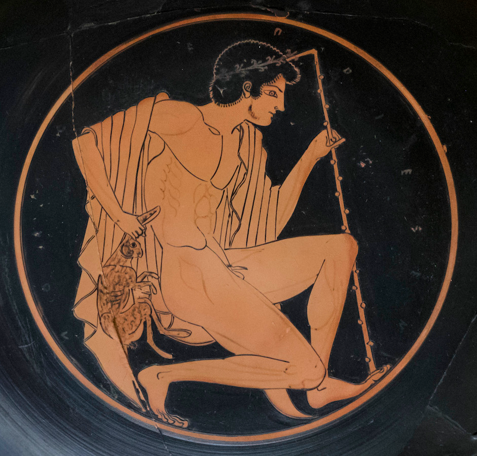 Ancient Greek red-figure pottery painting depicting a seated male figure holding a rod and a small hare, hanging from his left hand. The circular composition features detailed musculature and drapery, with Greek letters around the border indicating possible identification or context.