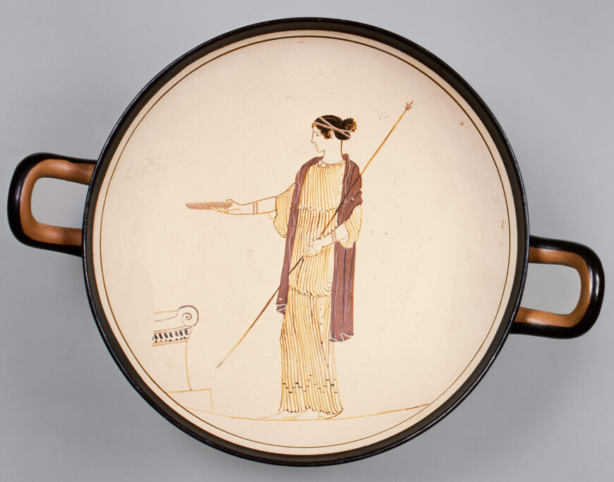 Photograph of an ancient Greek terracotta kylix (drinking cup) featuring a painted figure of a woman in profile holding a spear and extending her hand toward a small Ionic column fragment. The cup has two horizontal handles and a cream-colored interior with black and reddish-brown painted details.