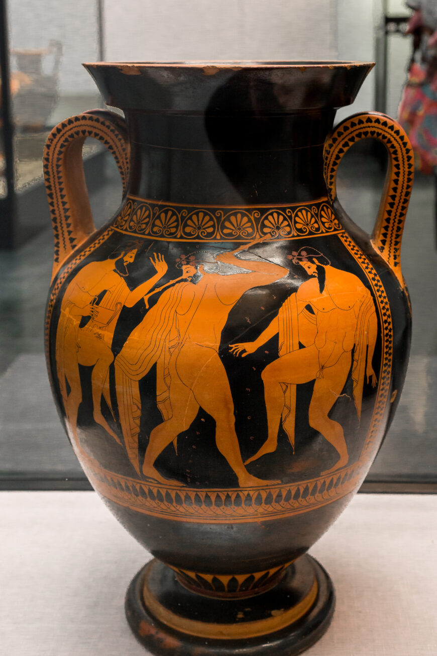 Photograph of an ancient Greek red-figure amphora depicting a mythological scene with three male figures. The vase features intricate floral patterns around the neck and handles, highlighting classical Greek pottery artistry.