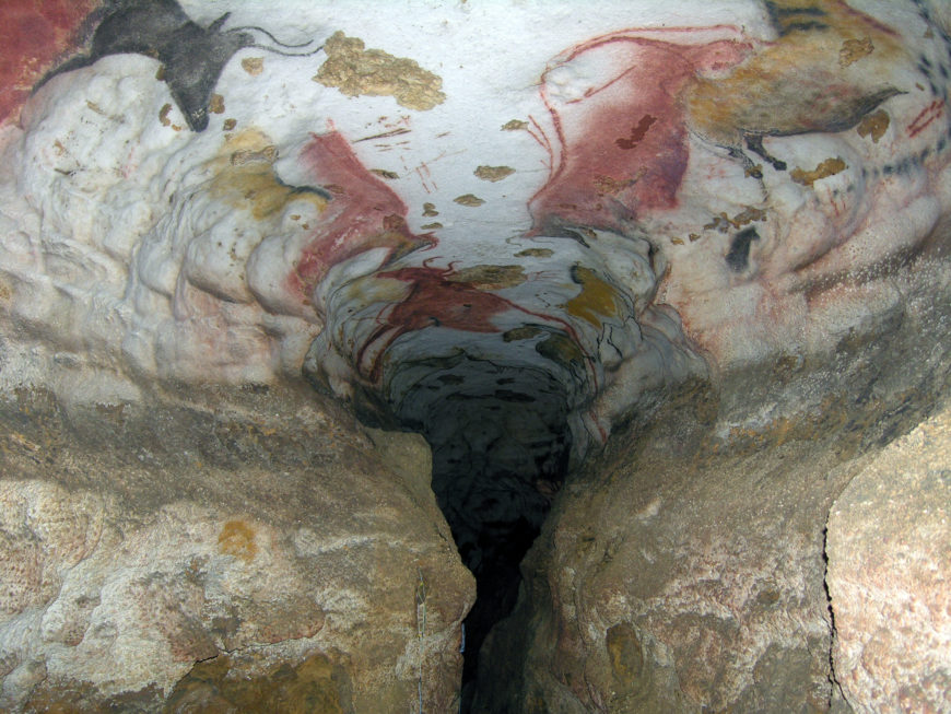Photograph of a cave interior featuring replicas of prehistoric wall paintings on the ceiling and upper walls. The artwork includes various animal figures in red, black, and yellow pigments, highlighting early human artistic expression.