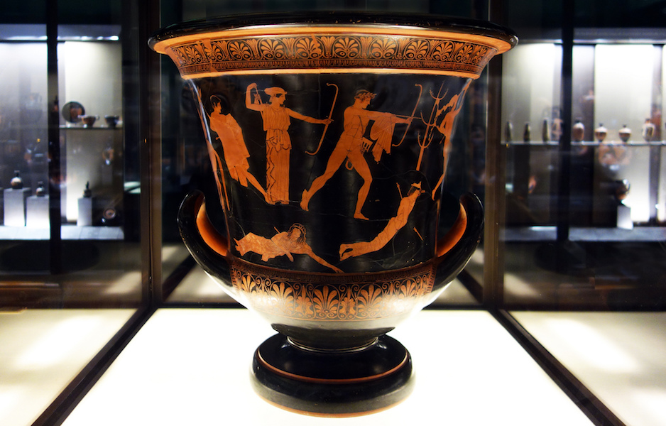 Photograph of an ancient Greek terracotta vase displayed in a glass case within a museum gallery. Vase features red-figure artwork depicting human and mythological figures, surrounded by other similar pottery pieces on illuminated shelves in the background.