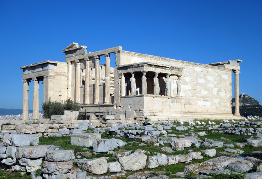 Photograph of the Erechtheion, an ancient Greek temple on the Acropolis of Athens, showcasing its iconic Caryatids—six draped female figures serving as architectural supports. The structure features white marble columns and walls, set against a clear blue sky with scattered ruins and greenery in the foreground.