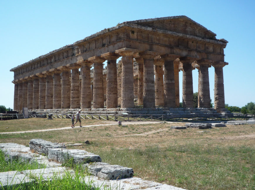 Photograph of an ancient Greek temple Hera II with Doric columns, showcasing classical architecture under clear blue sky. The structure features a rectangular layout with multiple columns on all sides, surrounded by grassy terrain and scattered stone ruins.