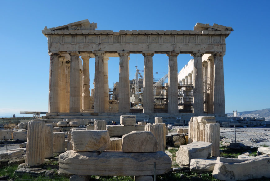Photograph of the Parthenon, an ancient Greek temple on the Acropolis of Athens, showcasing its iconic Doric columns and partially ruined structure. Scaffolding visible inside indicates ongoing restoration work, with scattered stone ruins in the foreground under a clear blue sky.
