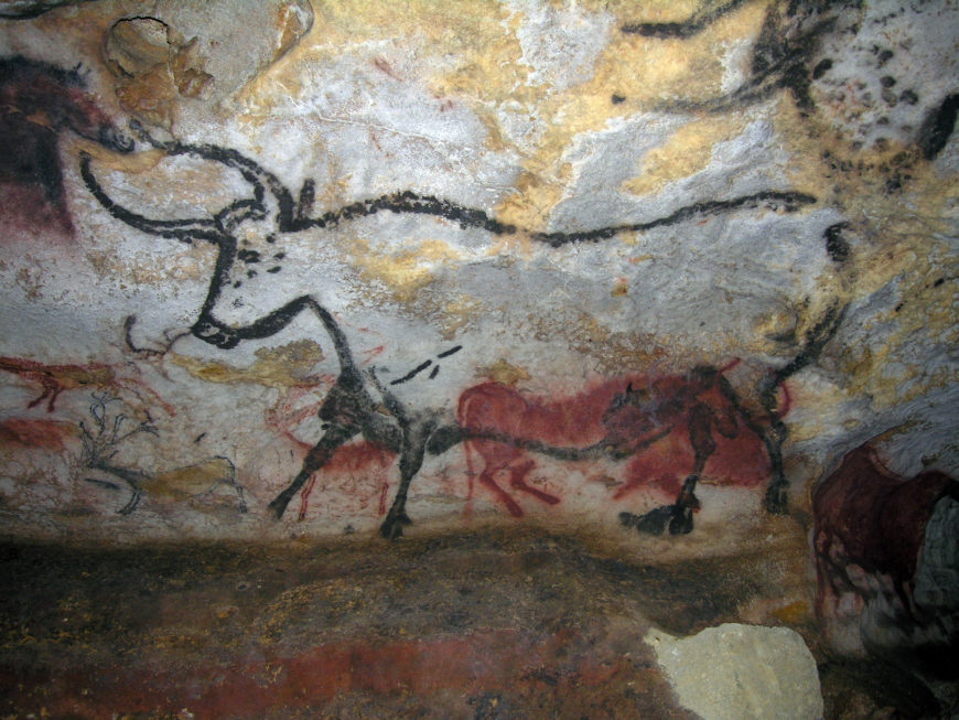 Photograph of a replica of prehistoric cave paintings depicting large animals, including a prominent bull with long horns and smaller figures in red and black pigments. The artwork features natural rock textures and showcases early human artistic expression.