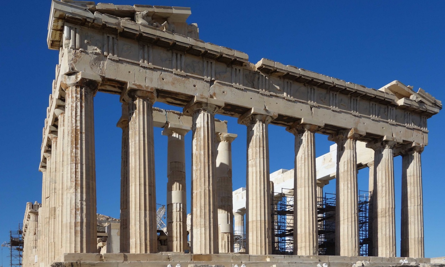 Photograph of Parthenon temple ruins in Athens, showcasing ancient Greek Doric columns and partially intact entablature under clear blue sky. Scaffolding visible inside structure indicates ongoing restoration efforts on this historic landmark.