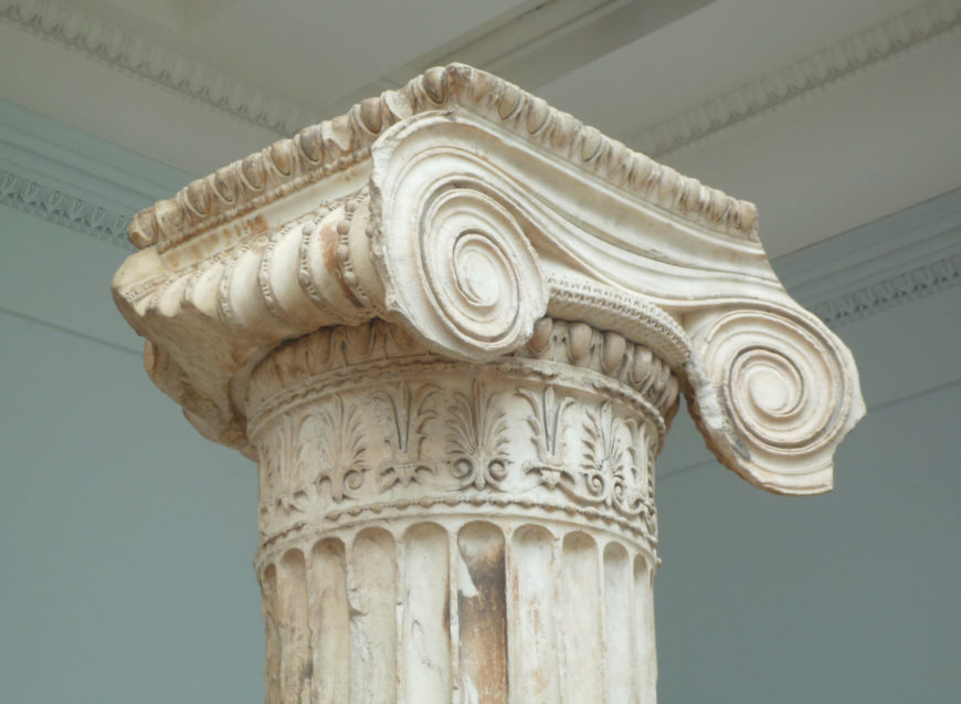 Photograph of a classical Ionic column capital from the North Porch of the Erechtheion showcasing detailed volutes and decorative floral patterns. The column features fluted shaft and intricate carvings, highlighting ancient Greek architectural design.