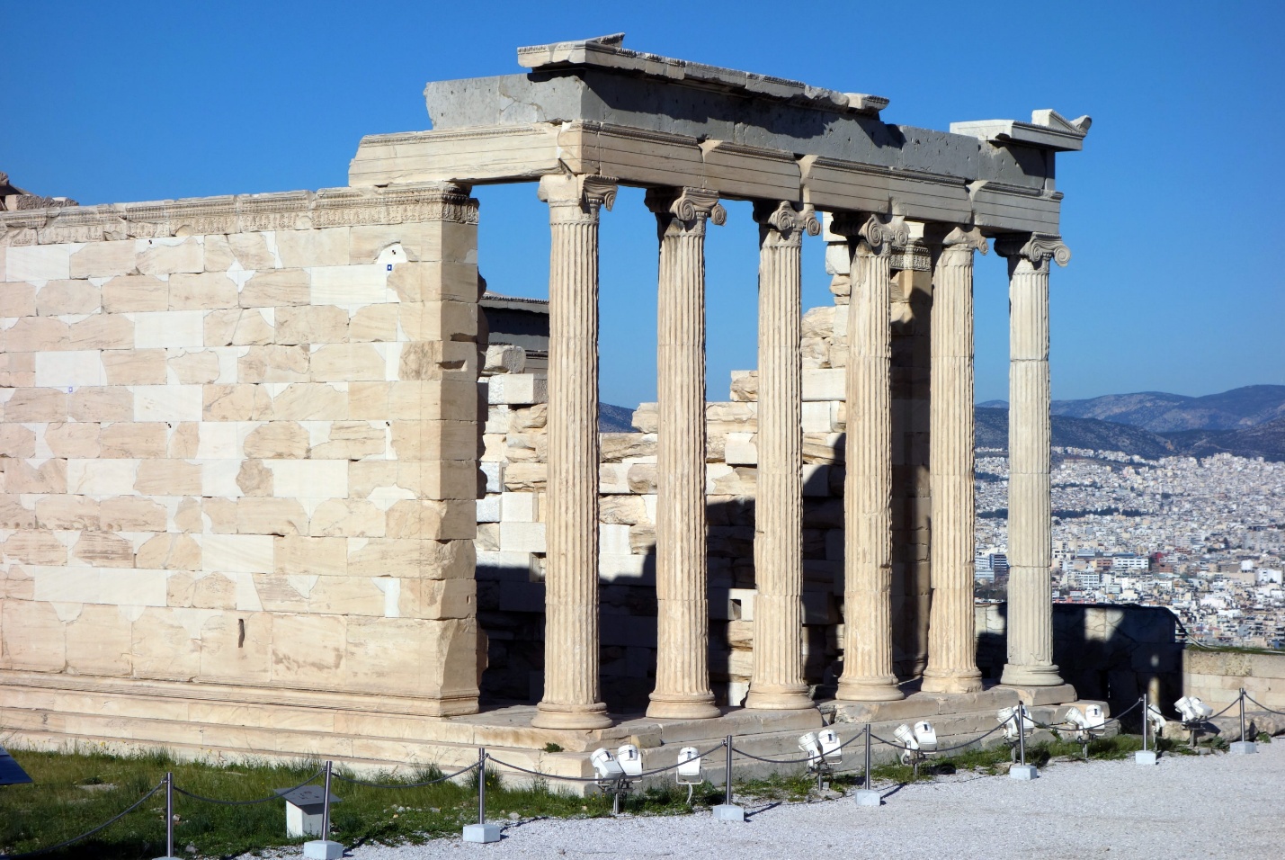 Photograph of East porch of the Erechtheion featuring six tall, fluted columns with Ionic capitals and a partially intact entablature. Background shows a sprawling cityscape under a clear blue sky, highlighting contrast between historic architecture and modern urban environment.
