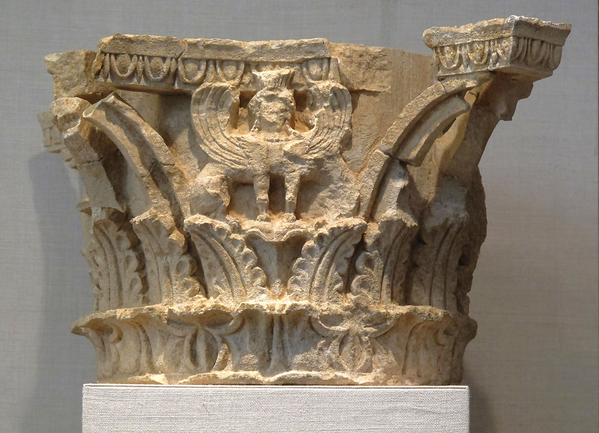 Photograph of Corinthian column capital featuring intricate carvings, including a central winged figure resembling a griffin or mythical creature. The capital displays symmetrical floral and geometric patterns with a weathered, beige surface.