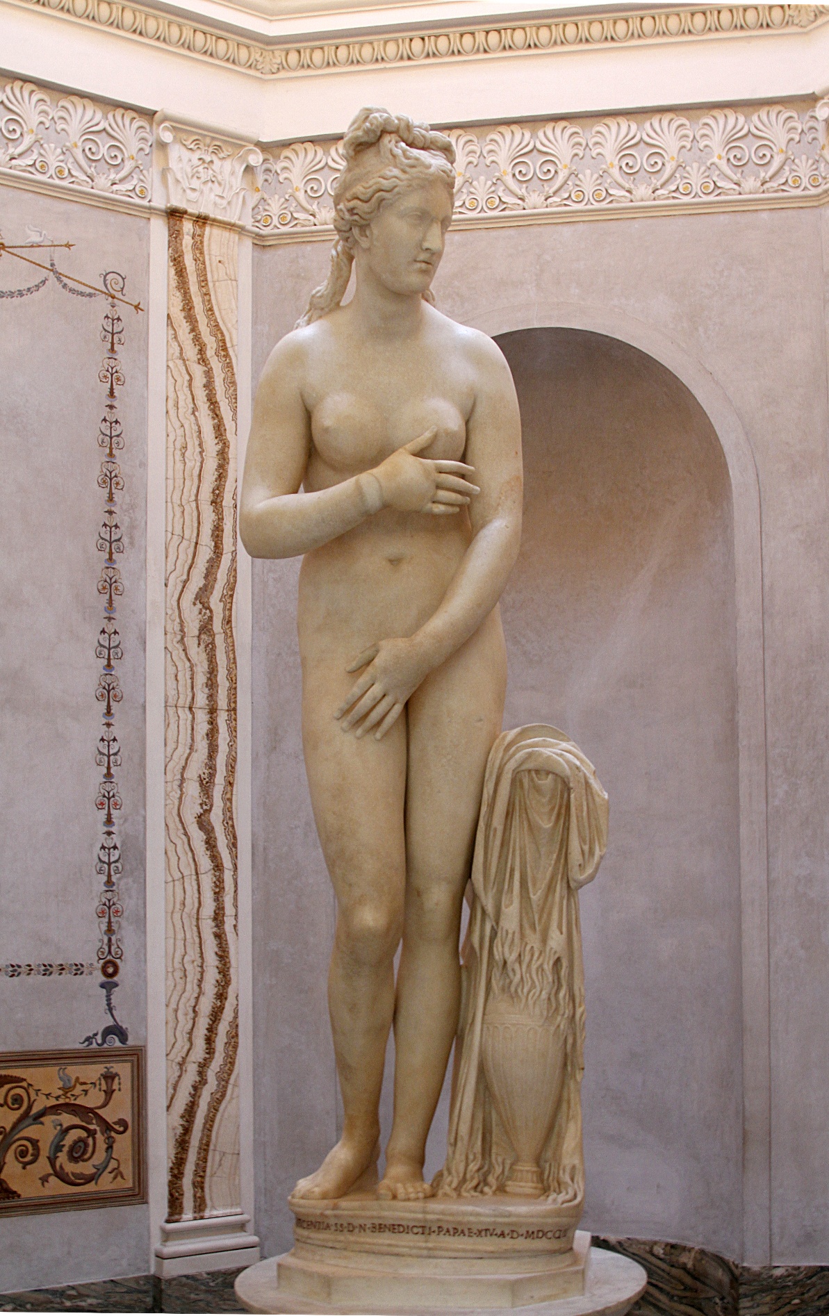 Capitoline Venus three-dimensional sculpture in white marble showing female figure in contrapposto stance next to a water vessel covered by a cloth. The figure is attempting to cover her private areas while looking to the side.