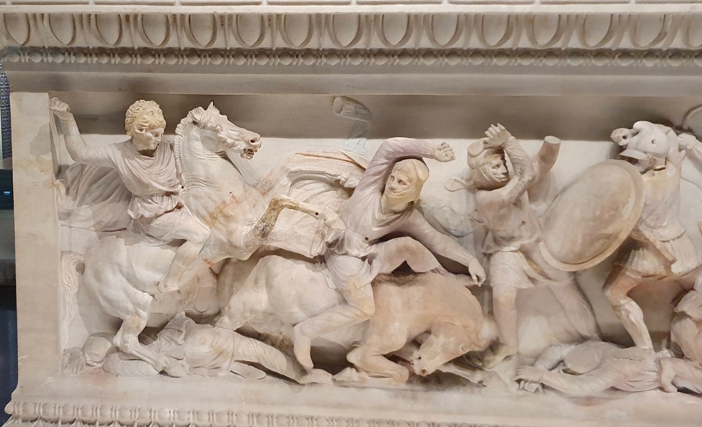 A photograph of an ancient marble relief sculpture depicting a dynamic battle scene with warriors, horses, and a fallen animal. The relief features detailed carvings of figures in motion, including a mounted soldier wielding a weapon, a shield-bearing warrior, and a charging bull, all framed by decorative borders.