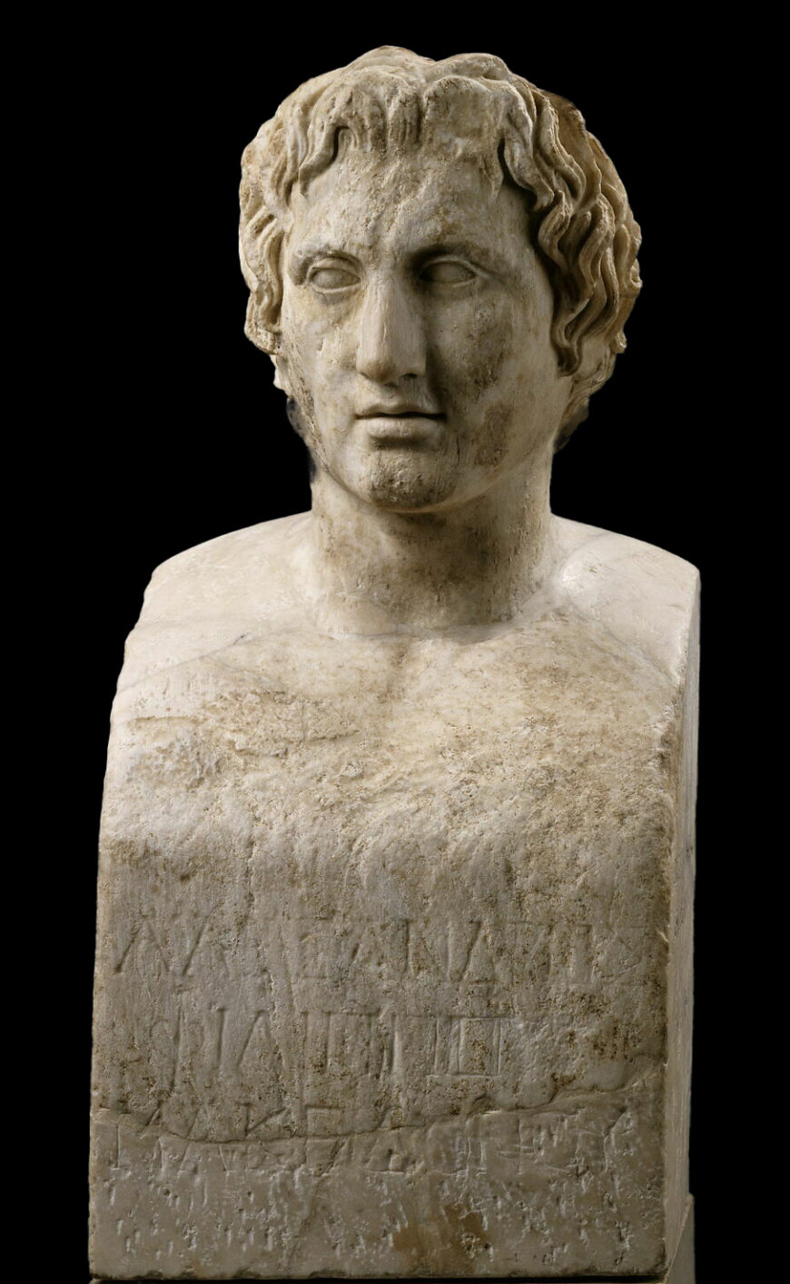 Photograph of Portrait of Alexander the Great marble bust with face, featuring curly hair and a rectangular base inscribed with worn Latin text.