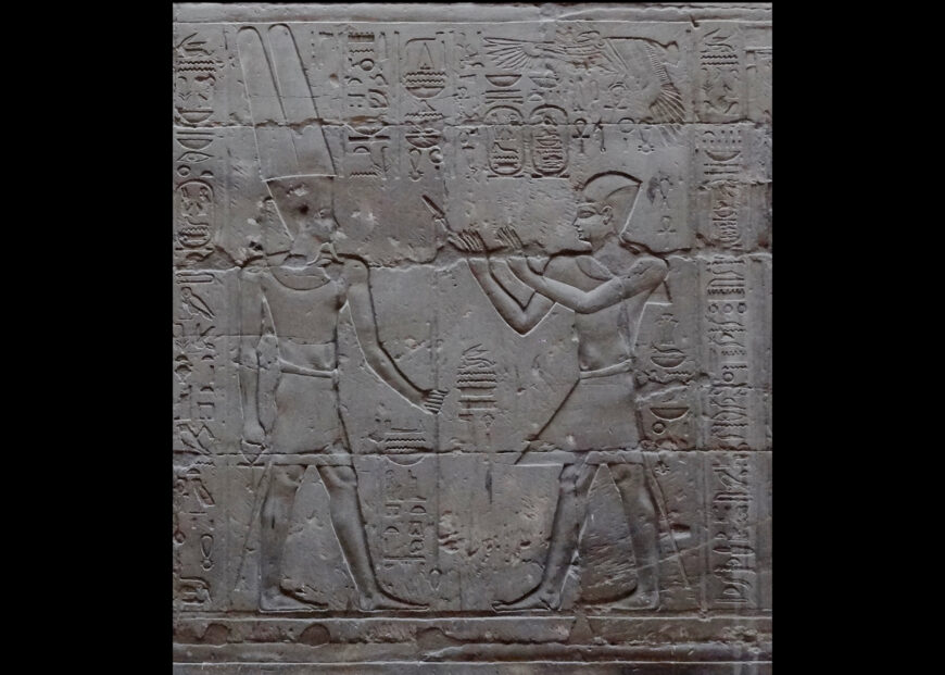 Bas-relief carving depicting two ancient Egyptian figures facing each other, one of which is Alexander the Great, surrounded by hieroglyphic inscriptions. One figure wears a tall crown and holds a staff.