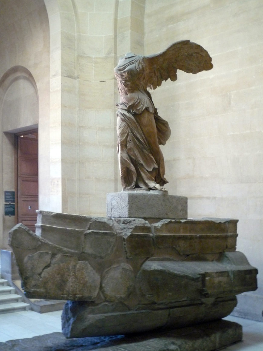Photograph of the Winged Victory of Samothrace, a marble statue of the Greek goddess Nike, displayed on a stone pedestal inside a museum with beige walls and an arched doorway. The statue features detailed drapery and large wings but lacks a head and arms, symbolizing victory and movement.