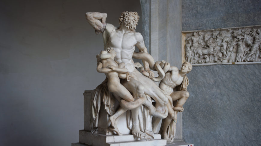 A photograph of the Laocoön and His Sons marble sculpture, depicting a muscular man and his two sons entangled by serpents. The sculpture highlights detailed anatomy, intense expressions, and dynamic poses, set against a stone wall with a carved relief panel.