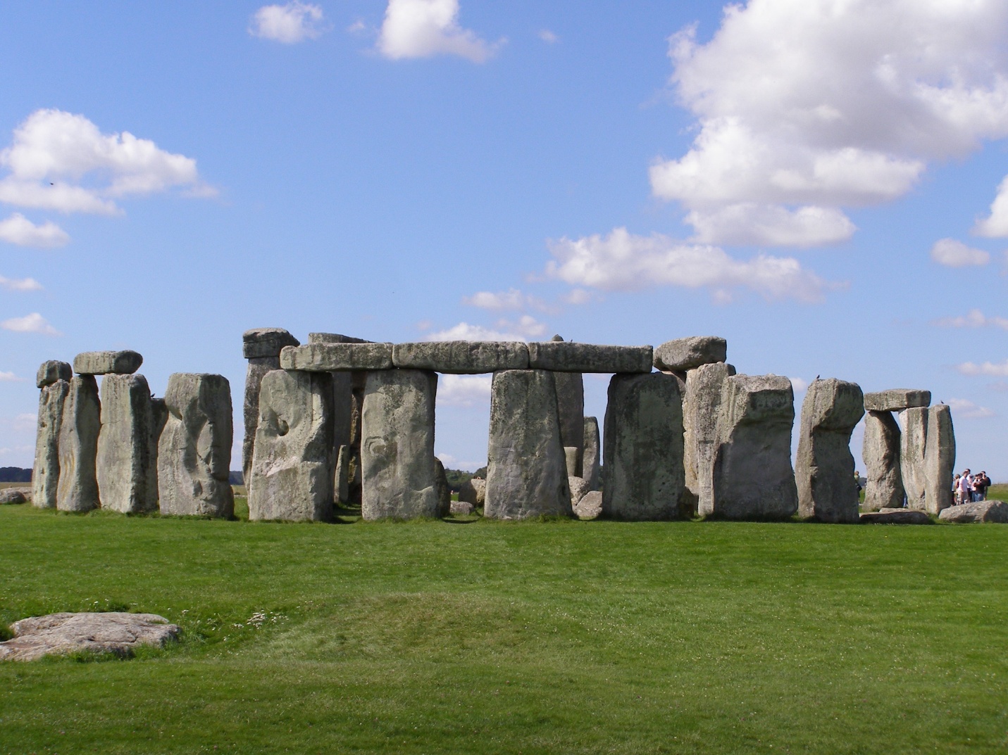 Photograph of Stonehenge, a prehistoric stone monument consisting of large standing stones arranged in a circular layout on a grassy plain under a partly cloudy sky. The image highlights the massive size and weathered texture of the stones, with some horizontal lintels resting atop vertical sarsen stones, illustrating the monument's iconic structure.