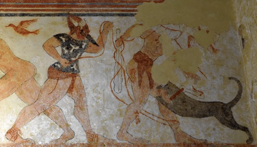 A photograph of an ancient wall painting depicting two human figures and a canine, with one figure holding a rope. The artwork features earthy tones, visible wear and damage.