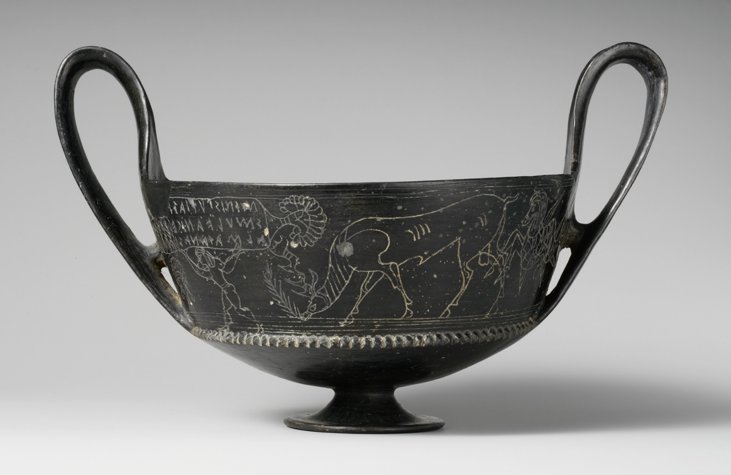 Photograph of an ancient Terracotta kantharos (vase), a shallow drinking cup with two handles and a footed base. The cup features detailed white line drawings depicting a stag and human figures, along with Greek inscriptions above the artwork.
