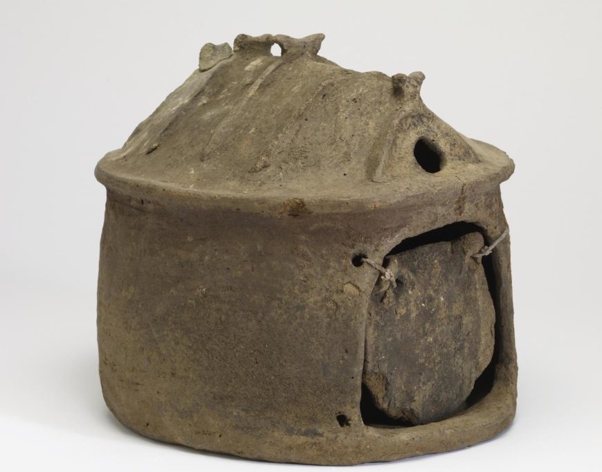 Photograph of a small, round, ancient clay hut urn with a domed top featuring ventilation holes and a front opening covered by a circular clay door tied with string. The piece shows signs of wear and has a rough, earthy texture.