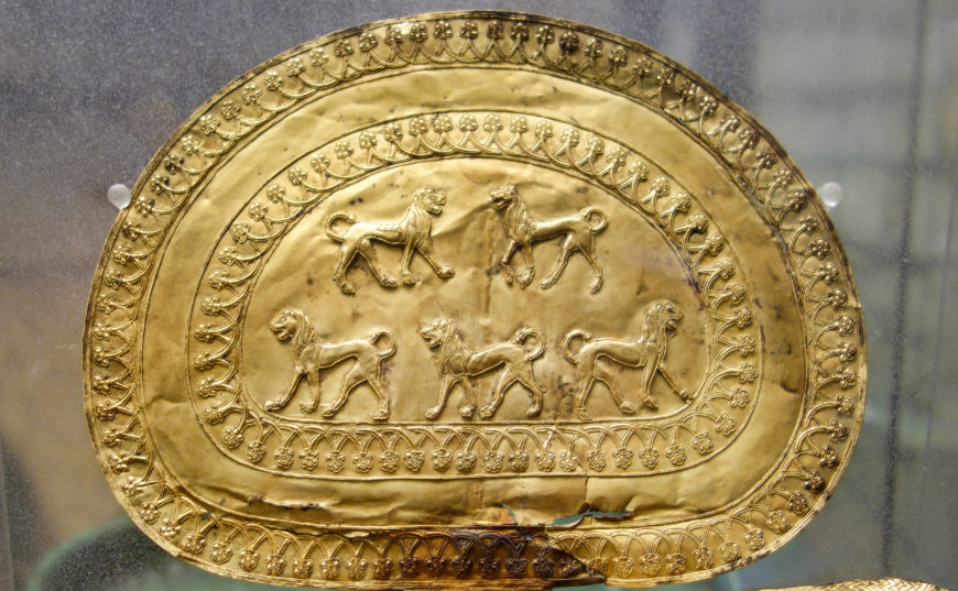 Photograph of an ancient gold Large Parade Fibula shaped like a rounded rectangle with intricate embossed designs. It features five stylized lions arranged in two rows, surrounded by decorative borders of floral and geometric patterns.