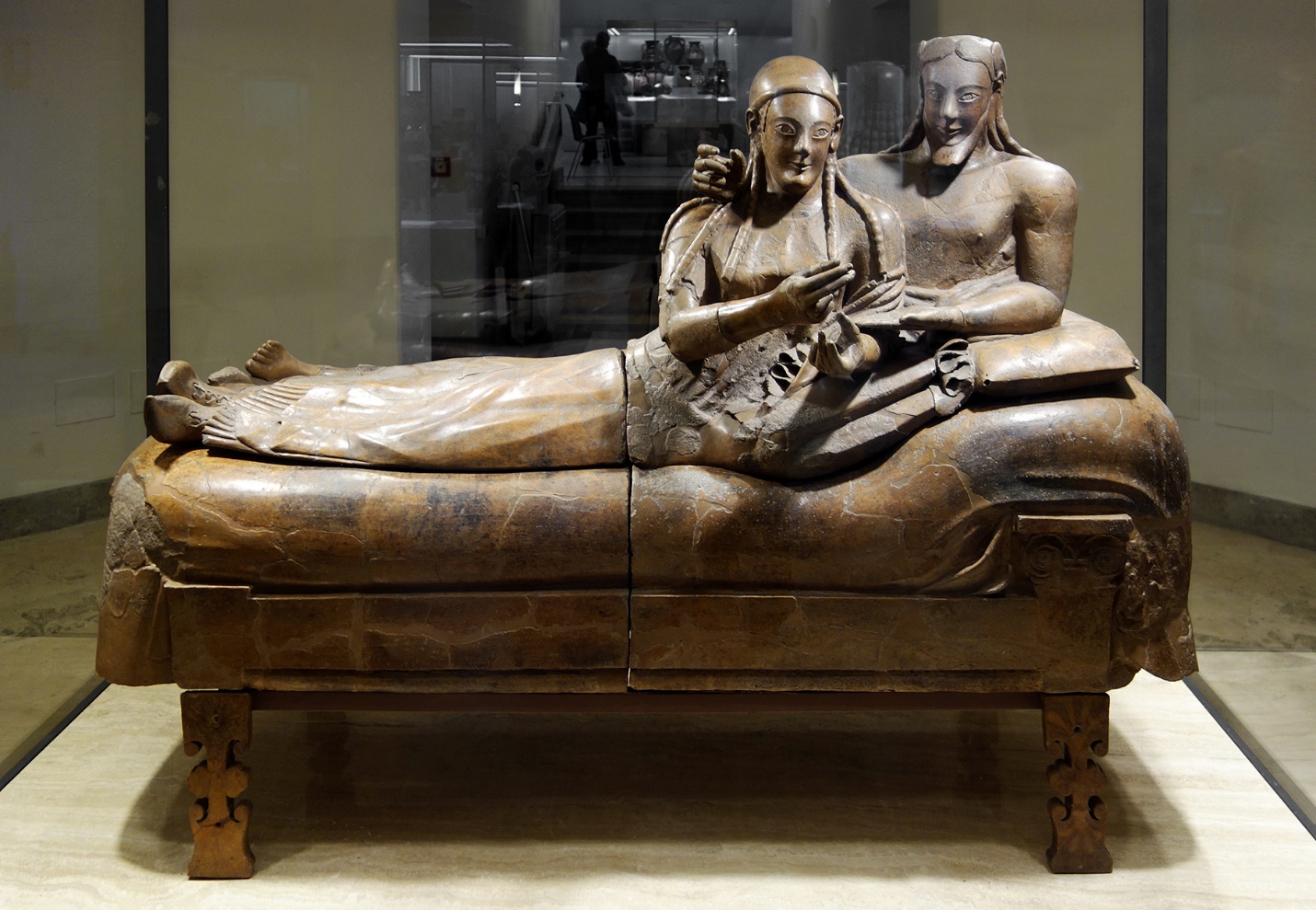Photograph of an ancient Etruscan sarcophagus depicting a reclining couple on a cushioned couch, showcasing detailed carvings and lifelike poses. The sculpture highlights cultural aspects of Etruscan funerary art, with the couple holding objects and resting on elaborately designed structure,, emphasizing intimacy and status.