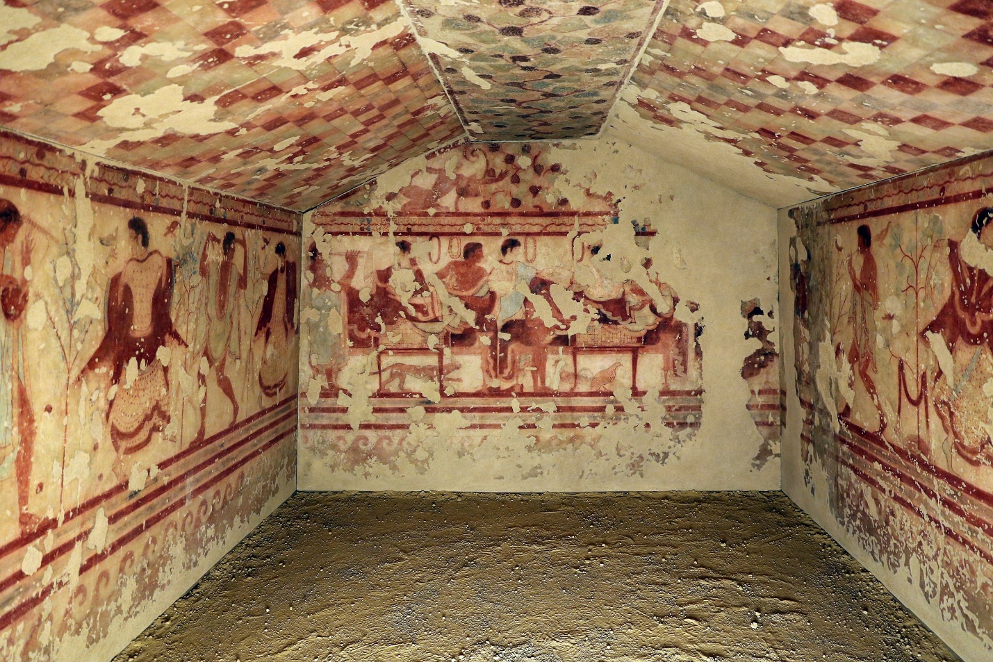 Photograph of an Tomb of the Triclinium interior featuring detailed frescoes on walls and ceiling depicting human figures in various poses and attire. The artwork uses red, brown, and beige tones with geometric patterns on the ceiling and narrative scenes on walls, highlighting cultural and ceremonial significance.