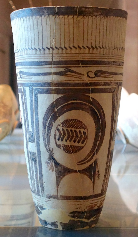 Photograph of an ancient ceramic pot featuring intricate black geometric and symbolic designs on a beige background. The cup displays a central circular motif with a patterned element inside, bordered by vertical and horizontal linear patterns near the rim and base.