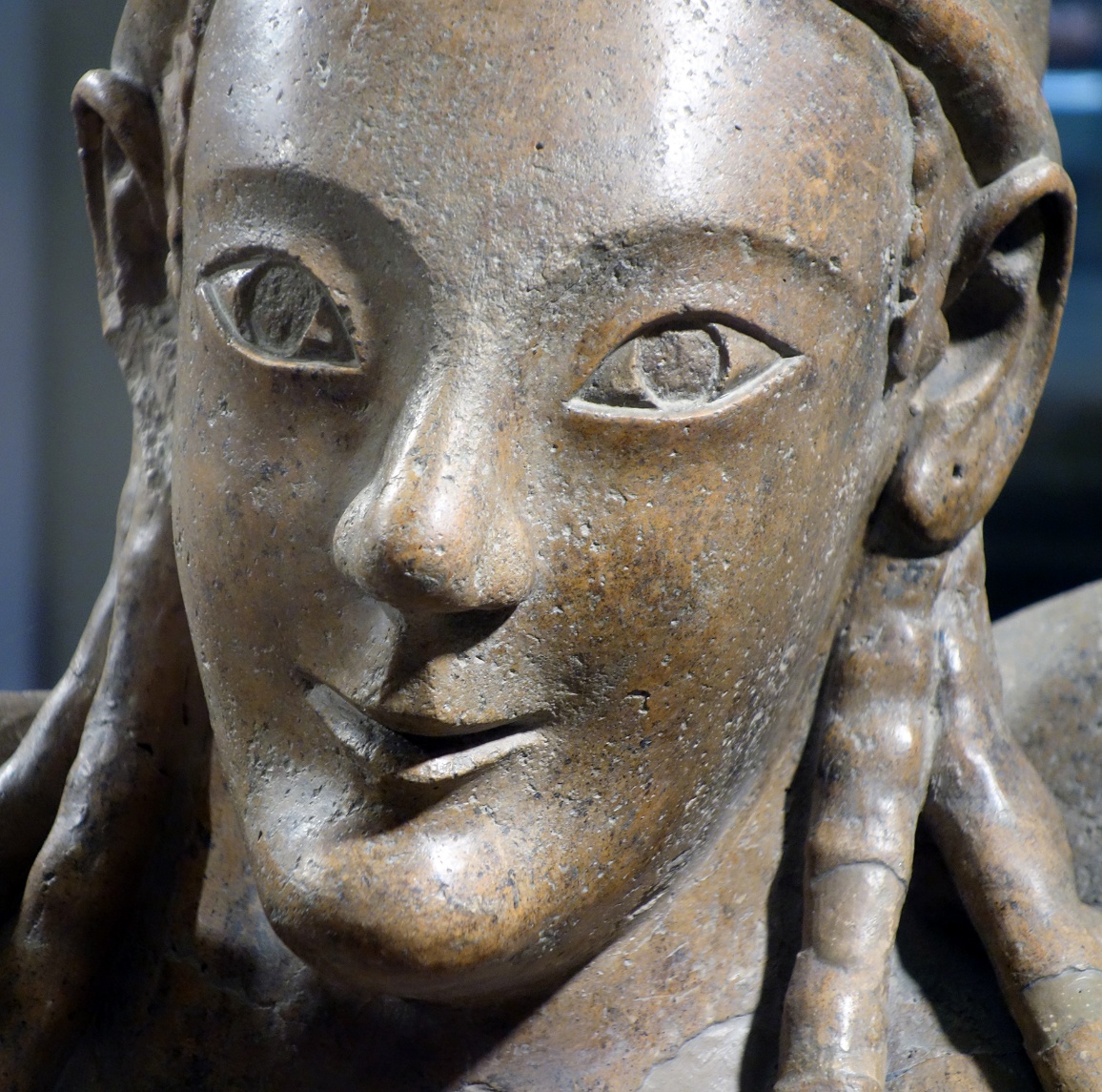 Close-up photograph Female figure's face (detail), Sarcophagus of the Spouses, terracotta sculpture depicting a human face with stylized almond-shaped eyes and a subtle smile. The sculpture features detailed hair strands and a smooth textured surface.