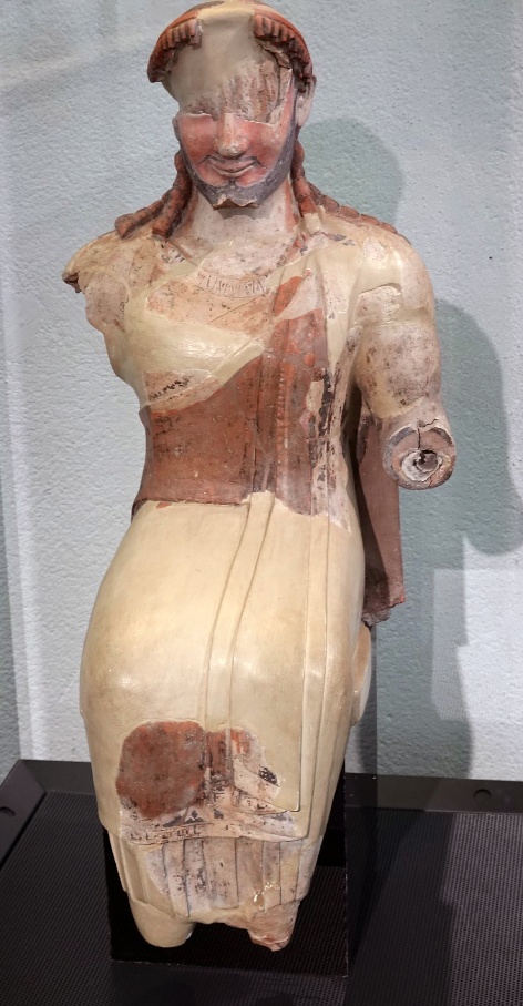 A photograph of an ancient marble statue depicting a seated man wearing a draped garment with traces of red and brown paint. The statue is missing its right arm and parts of the face, showcasing weathering and restoration efforts on a black pedestal against a plain background.