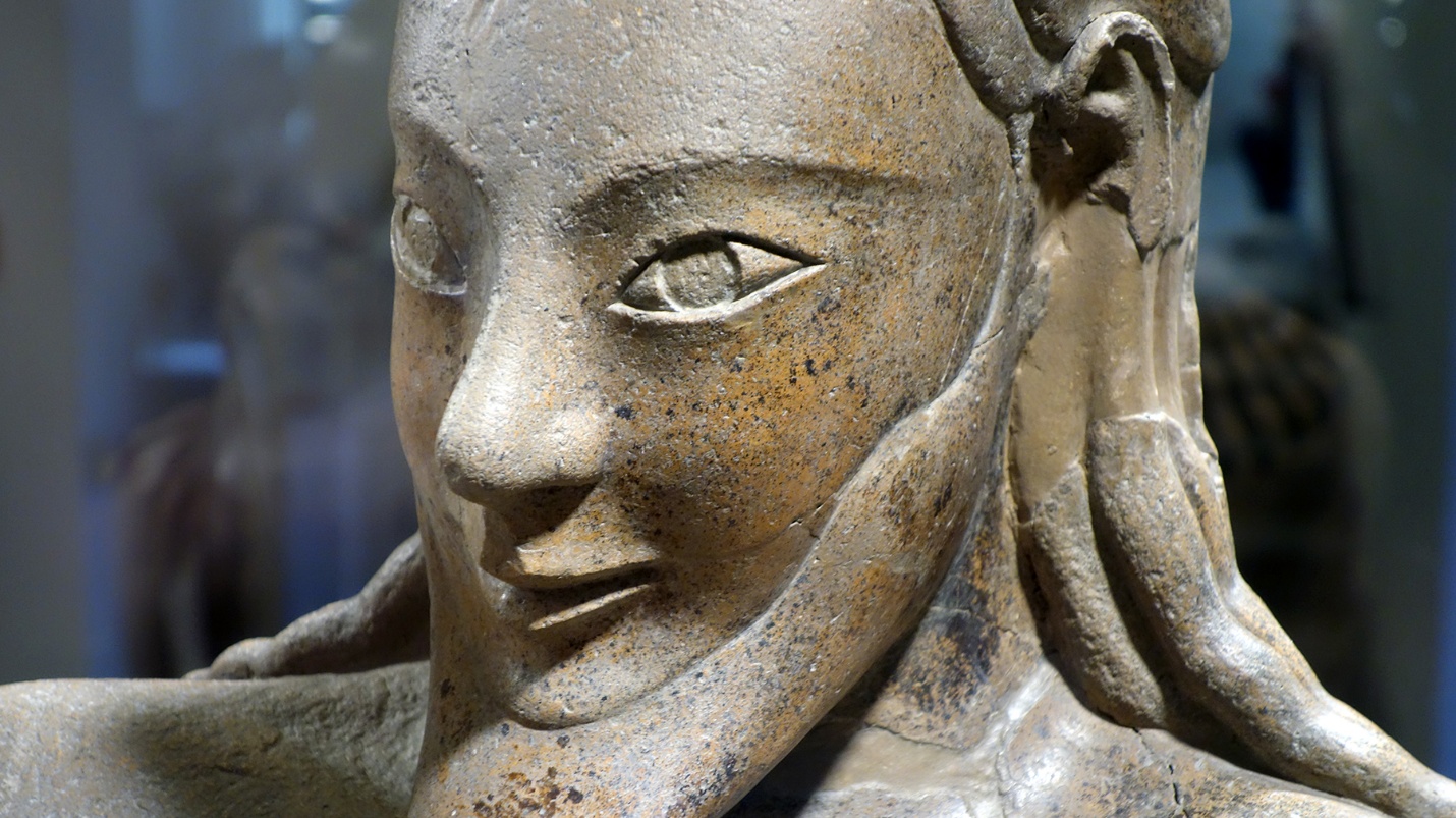 Male figure's face (detail), Sarcophagus of the Spouses depicting a seated figure with detailed carving on the face including a beard and almond shaped eyes. The sculpture appears weathered, showcasing texture and craftsmanship, with a blurred background suggesting a museum or display setting.