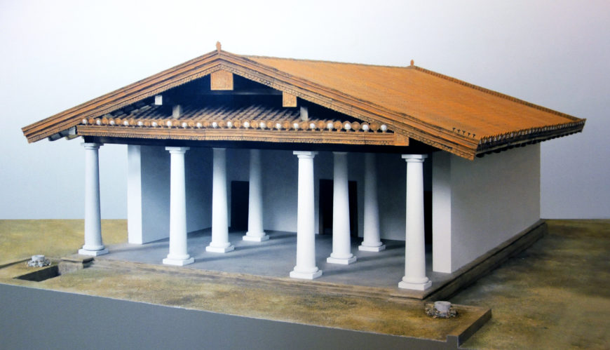 A photograph of a Reconstruction of an Etruscan Temple with a rectangular base and six white columns supporting a triangular wooden roof. The model highlights architectural features such as column spacing, roof beams, and decorative elements along the roofline.