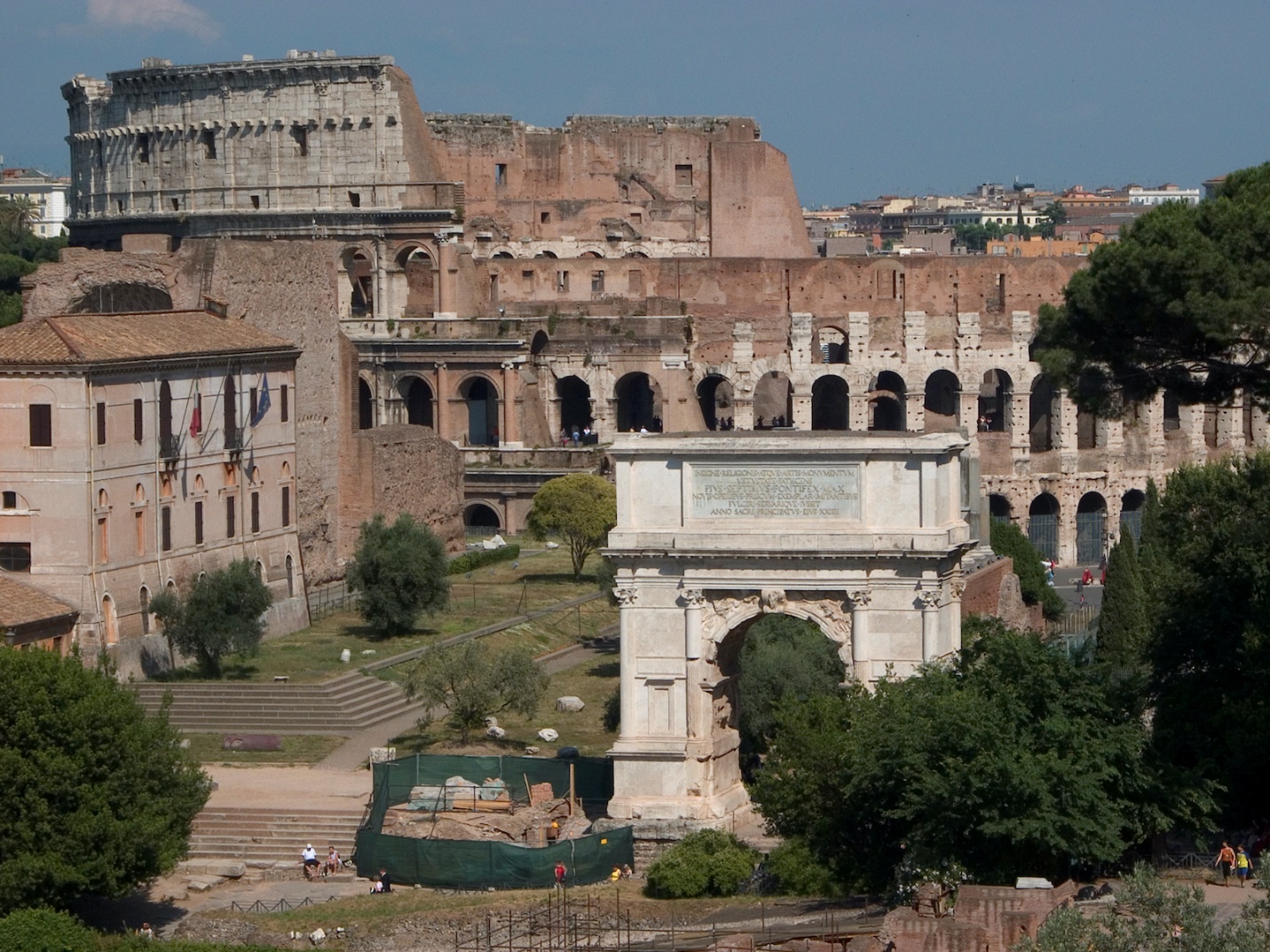 Photograph of ancient Roman ruins at the Roman forum featuring the Colosseum and Arch of Constantine under clear blue sky. The scene includes surrounding greenery, historic buildings, and pathways, highlighting architectural details and preservation state.