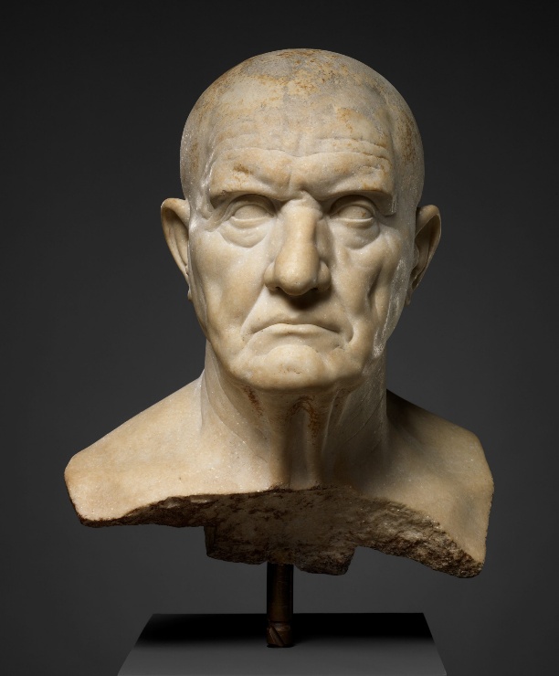 A photograph of an ancient marble bust depicting a male figure with a bald head and detailed ears, mounted on a stand. The bust shows signs of age with surface discoloration and a broken base