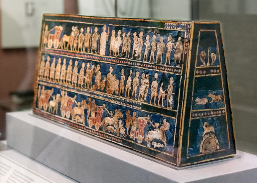 Photograph of an ancient trapezoidal artifact decorated with three horizontal registers of detailed human and animal figures in relief. The artifact features vibrant blue and gold colors, depicting scenes of daily life and ceremonial events, with additional smaller animal motifs on the side panels.
