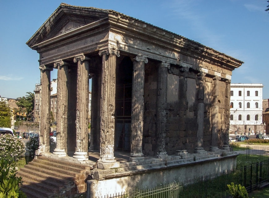 Photograph of an ancient Roman temple of Portunus featuring tall Ionic columns and a triangular pediment, showcasing classical architectural design. The structure is weathered with visible wear on stone surfaces, situated in an urban setting with modern buildings and greenery in the background.