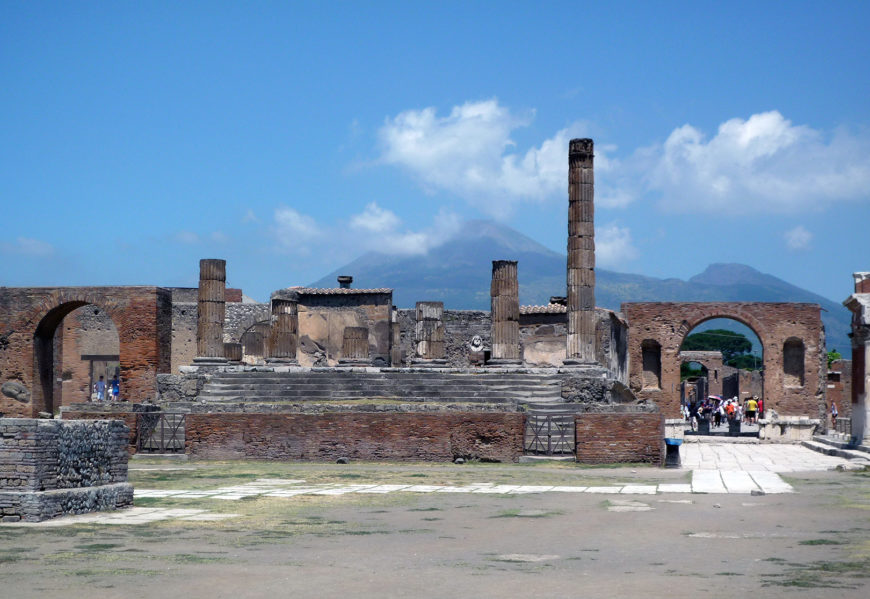 Photograph of Forum, Pompeii, looking toward Mt. Vesuvius with stone and brick structures, including columns and arches, under a bright blue sky with scattered clouds. Mount Vesuvius is visible in the background, highlighting historical significance and volcanic landscape near Pompeii.