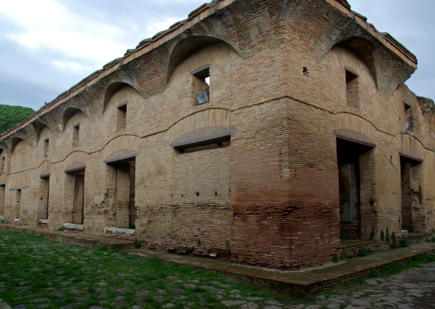 Photograph of an ancient brick structure with multiple arched windows and doorways, showcasing Roman architectural style. The building features weathered bricks with a distinct horizontal color variation and is set against a cloudy sky with green grass and stone pavement surrounding it.