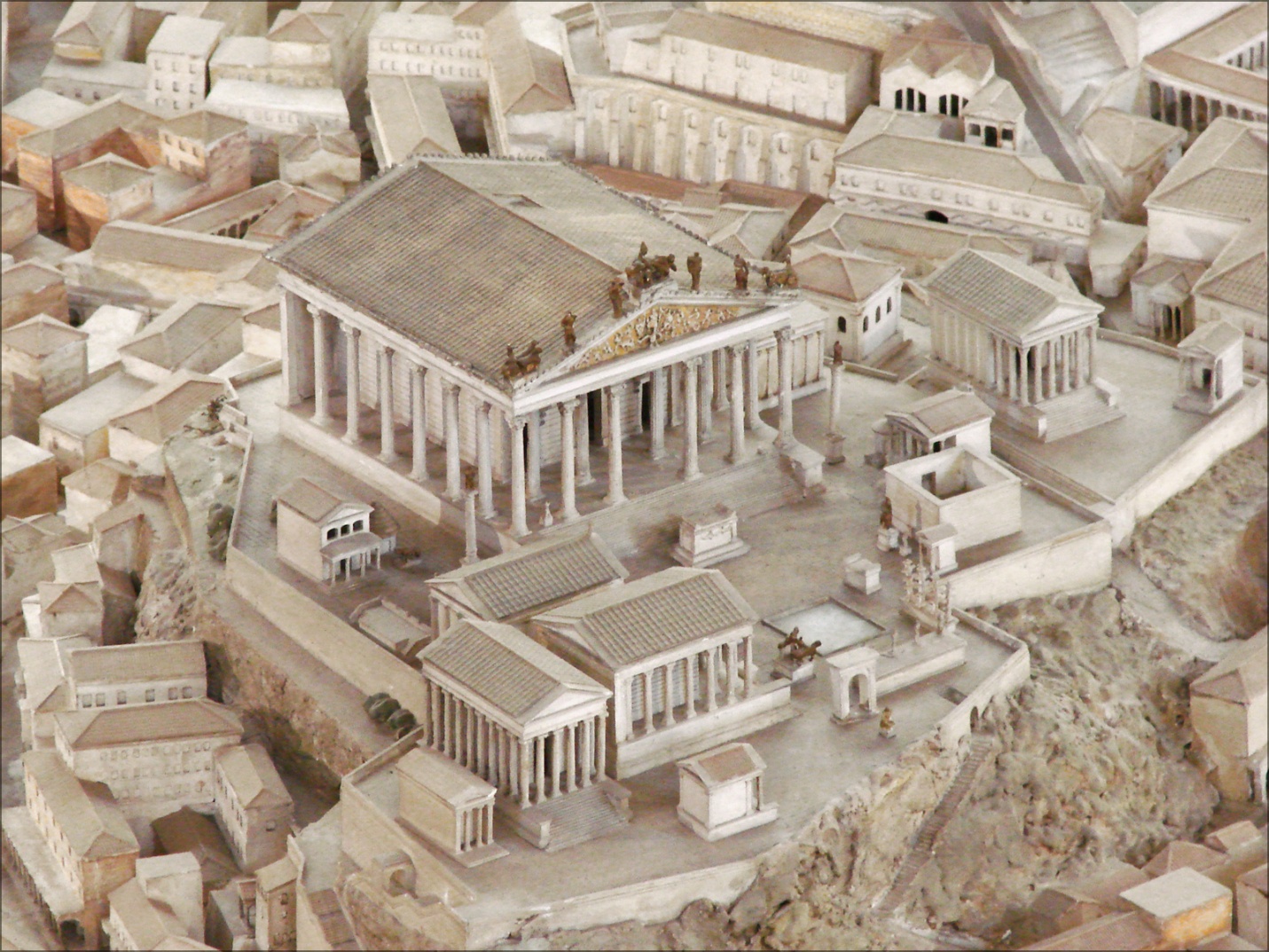A detailed architectural model depicting Temple of Jupiter Optimus Maximus, Capitoline Hill with multiple classical buildings featuring columns, pediments, and statues. The model highlights key structures such as a large temple with a triangular pediment adorned with sculptures, surrounded by smaller temples and public buildings arranged on a raised platform.