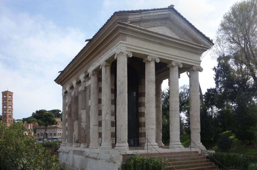 Photograph of an Temple of Portunus featuring tall, fluted Ionic columns supporting a triangular pediment. Surrounding greenery and a distant bell tower provide historical and architectural context.