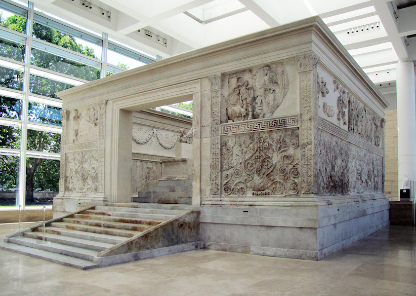 Photograph of Altar of Augustan Peace large marble structure with intricate bas-relief carvings depicting figures and decorative patterns. The structure features a wide entrance with steps leading up and is displayed indoors with large windows allowing natural light.