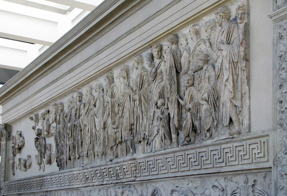 Photograph of processional scene (south side), Ara Pacis Augustae (Altar of Augustan Peace) ancient marble relief panel depicting a procession of robed figures, including men, women, and children, carved in high detail. The panel features intricate patterns along the bottom edge and is displayed indoors with natural light illuminating the sculpted figures.