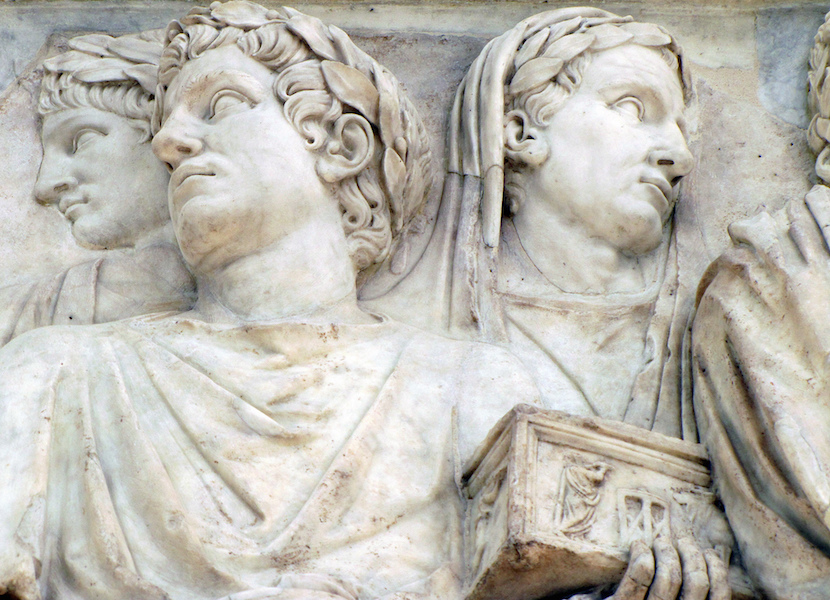 A photograph of an ancient marble relief sculpture depicting two robed figures with laurel wreaths, one holding a detailed incense box. The sculpture features intricate carvings of facial expressions, clothing folds, and symbolic elements.