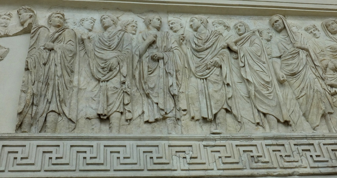 A photograph of an ancient stone relief sculpture depicting a group of robed figures engaged in conversation or interaction. The relief features detailed carvings of clothing folds and facial expressions, with a decorative geometric border along the bottom.