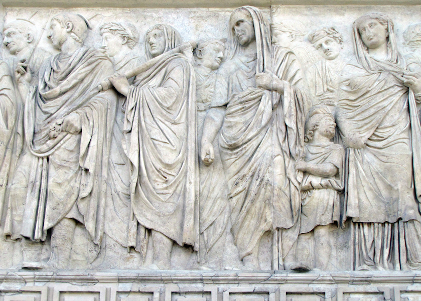 A marble relief sculpture depicting a group of robed figures in classical attire, arranged in a procession. The artwork features detailed drapery, varied poses, and expressions, highlighting ancient Roman artistic style and cultural themes.