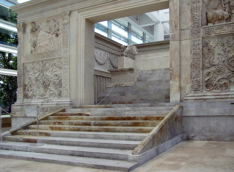 Photograph of detail of Altar of Augustan Peace large marble structure with intricate bas-relief carvings depicting figures and decorative patterns. The structure features a wide entrance with steps leading up and is displayed indoors with large windows allowing natural light.