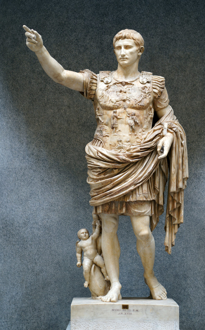 A photograph of Augustus of Primaporta marble statue depicting a Roman military leader in detailed armor and draped cloth, standing with one arm extended and the other holding a staff. A small cherub-like figure is positioned at the leader's right leg, and the statue rests on a rectangular pedestal with an inscription.