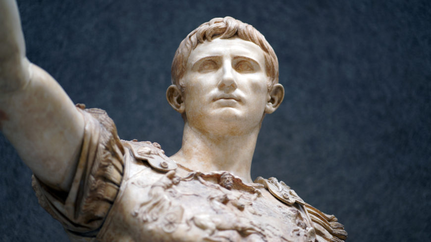 A photograph of Detail, Augustus of Primaporta marble statue depicting a figure in Roman military attire with an outstretched arm. The statue features detailed armor with ornate decorations and a textured background.
