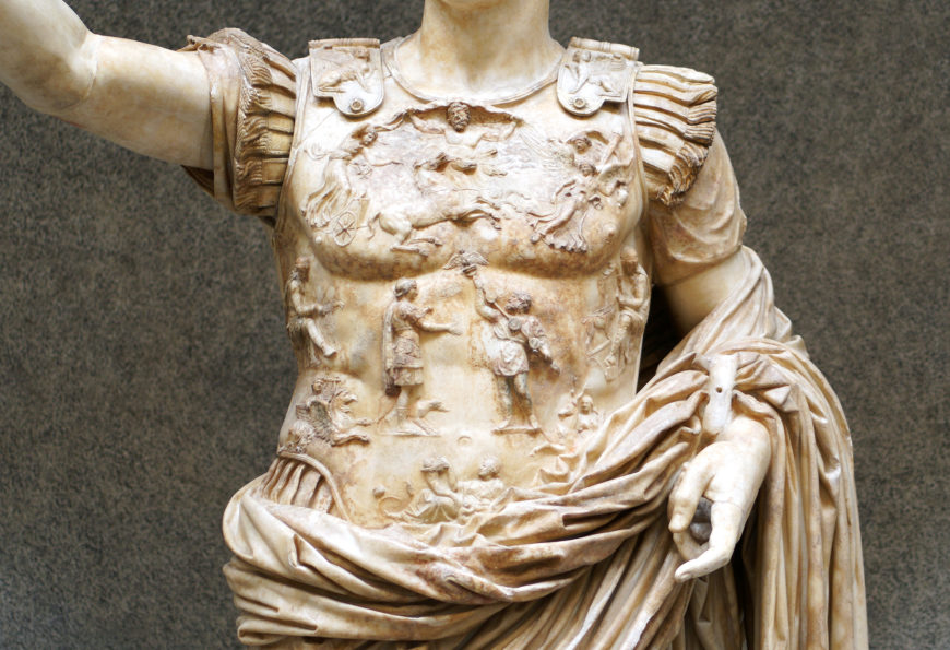 Photograph of Detail of the breastplate, Augustus of Primaporta marble statue torso depicting a Roman soldier wearing detailed armor with intricate relief carvings of mythological and battle scenes. The statue features a draped cloth around the waist and finely carved shoulder guards, highlighting craftsmanship and historical Roman military attire.