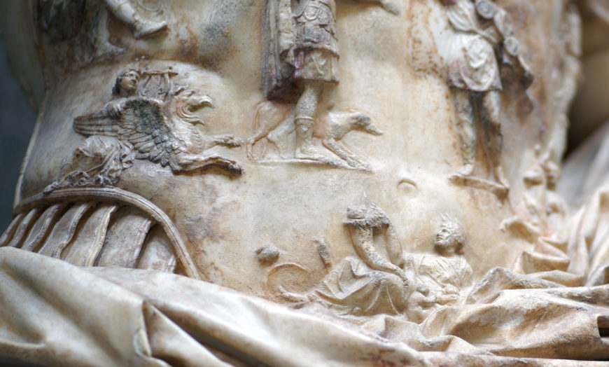 Close-up photograph of an ancient marble sculpture featuring detailed relief carvings of mythological figures, including a winged creature and a reclining human figure. The sculpture displays intricate textures and folds in the stone, highlighting craftsmanship and storytelling elements from classical antiquity.