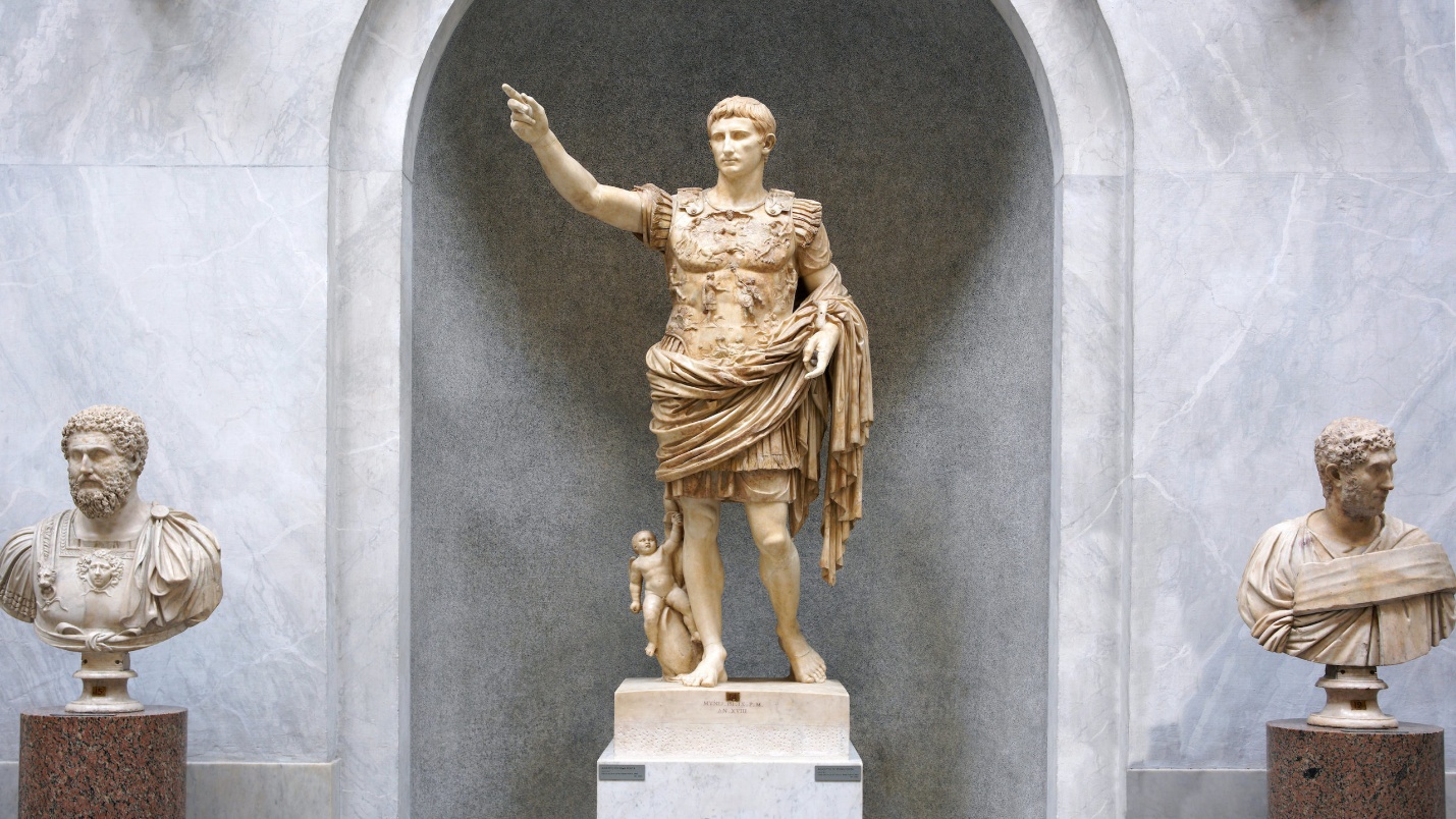 Photograph of three classical marble sculptures displayed against a marble wall with an arched niche. Central statue depicts a Roman figure in military attire with an outstretched arm and a small child at his side, flanked by two busts of similarly dressed figures on pedestals.